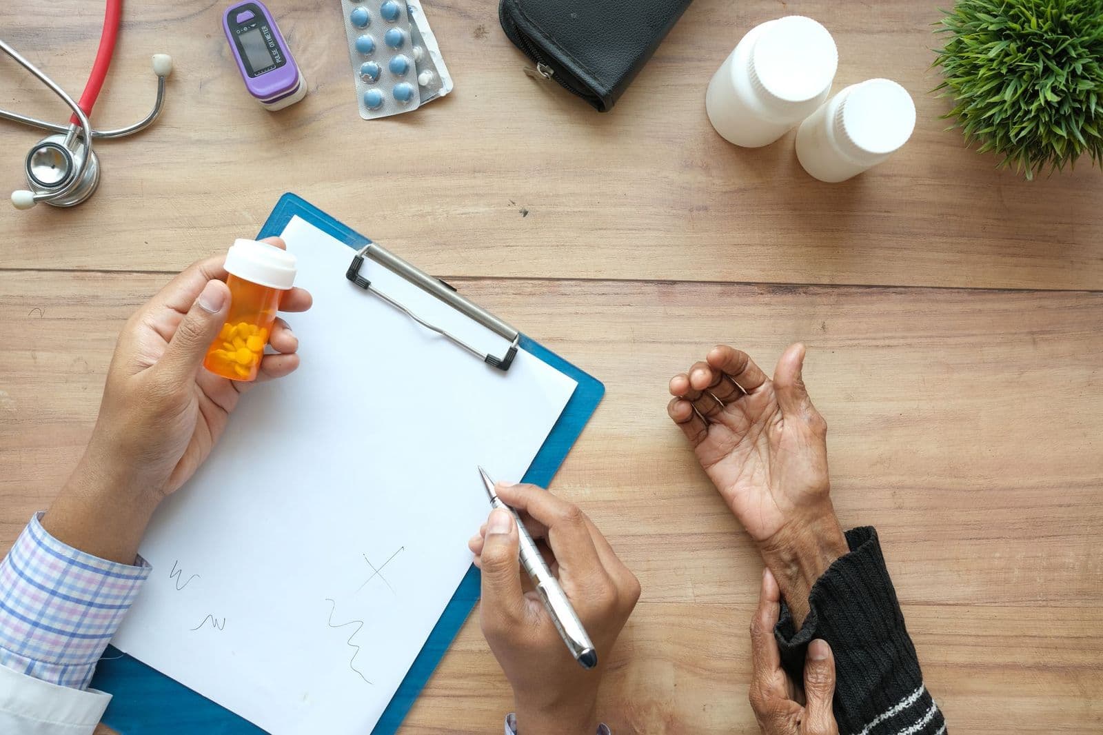 Vista superior da mesa de consultório com medicações, prancheta e as mãos do médico e de um paciente idoso