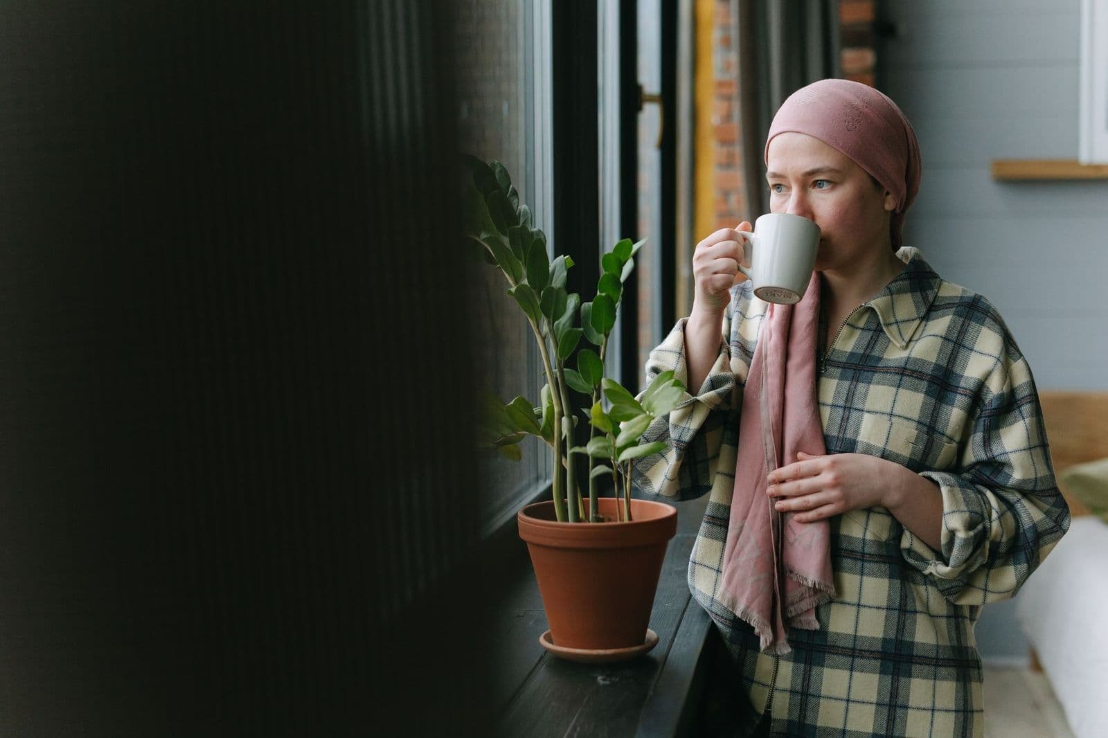 Mulher com lenço na cabeça tomando café junto à janela durante o tratamento oncológico